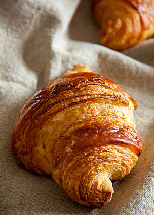 Ein goldbraun glänzendes Croissant mit sichtbaren Schichten liegt auf einem hellbeigen Leinentuch.