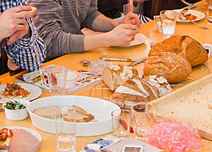 Eine kleine Gruppe von Menschen bei einer gemeinsamen Mahlzeit am Tisch. Im Vordergrund erkennbar frisches Brot nebst leckerem Belag und frischen Lebensmitteln.