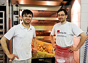 Bäcker Ricardo Fischer und Lutz Geißler stehen vor einem Bäckerofen und einem Korb frischer Brötchen.
