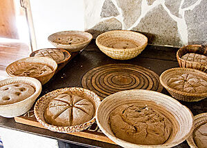 Auf der Oberfläche kunstvoll verzierte Brot-Teiglinge liegen in Gärkörben auf einem Tisch.