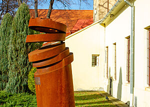 Auf einer Grünfläche neben einem Haus steht ein verrosteter Stahlzylinder, dessen oberes Drittel aussieht, als wäre es in Scheiben geschnitten. 