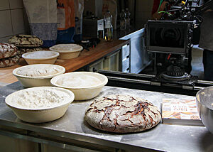 Mehrere Gärkörbe mit Teiglingen sowie fertig ausgebackene Brote liegen auf der Arbeitsfläche.