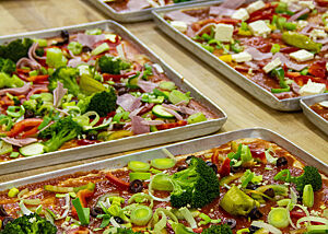 Mit Brokkoli, Lauch, Paprika und Tomatensoße belegte Pizzen liegen auf den Blechen.