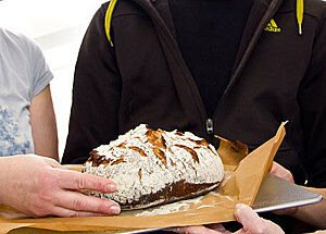 Eine Person reicht einer anderen Person ein frischgebackenes Brot auf einem Backblech. Die Kruste des Brotes ist bemehlt und zeigt eine rustikale Kruste.
