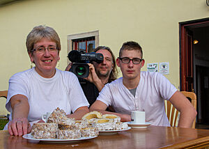 Martha und Lukas sitzen am mit Skalický Trdelník gedeckten Tisch, hinter ihnen hockt Kameramann Holger mit einer Filmkamera auf der Schulter.