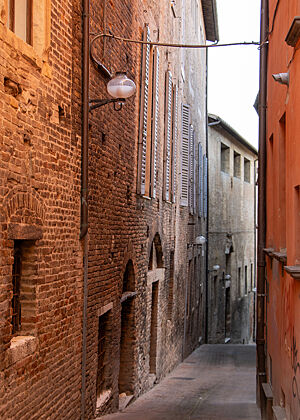 Historische Gasse in Urbino. Die Backsteinmauern der Gebäude links und rechts der schmalen Gasse ragen hoch hinauf und sind nur von kleinen Fenstern durchbrochen.