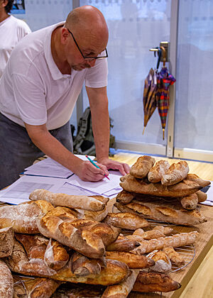 Der Baguette-Kurs ruft – Schelli schreibt noch schnell ein neues Rezept zum Experimentieren auf. An einem mit Baguettes bereits gut gefülltem Tisch entwickelt Schelli ein Rezept zum Experimentieren für den Baguette-Kurs.