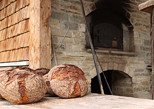 Neben einem Holzbackofen liegen zwei kräftig ausgebackene, rustikal aufgerissene Brotlaibe.