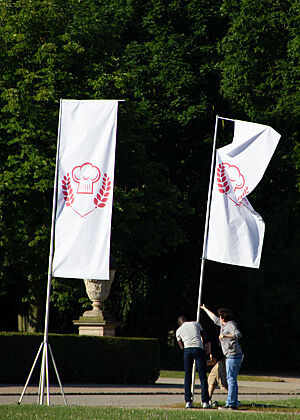 Fahnenbau für den Einmarsch der Jury und der Teilnehmer. Weiße Fahnen mit dem rotem Emblem der Sendung „Deutschlands bester Bäcker“ werden aufgestellt.