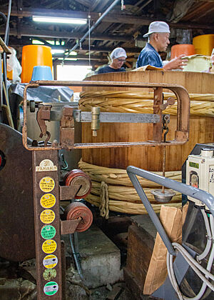 Holzfass zum Dämpfen von Reis Hinter einer verrosteten Waage steht ein großes Holzfass, dahinter der Chef des Betriebes und einn Teilnehmer der Reise.