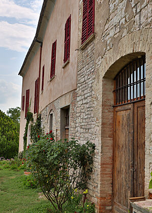 Die mit Rosenbüschen bepflanzte Fassade des Anwesens von Adriana Cucinelli ist teilweise hellrosa verputzt. Die Fensterläden sind dunkelrot gestrichen.