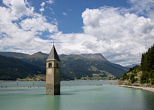 Aus dem türkisfarbigen Wasser des Reschensees ragt ein schlichter Kirchturm mit grauem Schieferdach auf. 