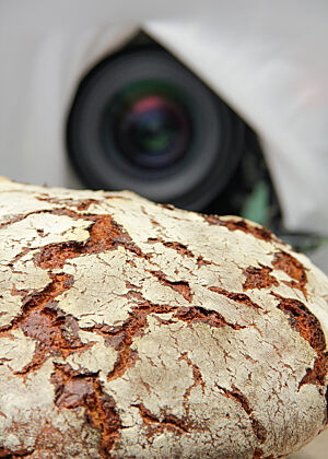 Das Brot im Mittelpunkt: Filmdreh in der Backstube. Ein rustikal aufgebrochenes und kräftig ausgebackenes Sauerteigbrot liegt vor einer Kamera.