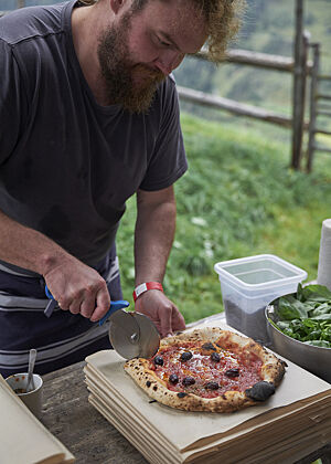 Eine Holzofenpizza, belegt mit Tomatensauce und schwarzen Oliven, wird mit einem Pizzarad geschnitten.