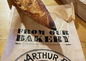 Zu Gast in der King Arthur Bakery. Ein rustikal ausgebackenes Baguette liegt auf einer Brottüte der King Arthur Bakery.