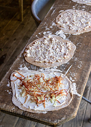 Mehrere Flammkuchen, dünn mit weißem Schmand bestrichen und mit goldbraunen Speckstücken bestreut, warten auf eine Holzbrett auf den Ofen.