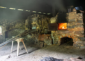 Die zwei baugleichen Holzöfen stehen nebeneinander in einer dunklen Scheune. Im linken Ofen ist dank der offenen Ofentüre das Leuchten des Feuers sichtbar.