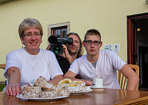 Kameramann Holger … Martha und Lukas sitzen am mit Skalický Trdelník gedeckten Tisch, hinter ihnen hockt Kameramann Holger mit einer Filmkamera auf der Schulter.