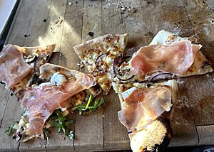 Pizzastücke einer Pizza Bianca liegen auf einem Holzbrett. Nach dem Backen wurden noch Rucola und Schinken auf die Pizzastücke gelegt.