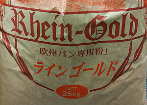 Ein Mehlsack tragt die altdeutsche Aufschrift „Rhein-Gold 25 kg“ sowie japanische Schriftzeichen.