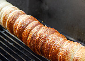Nur mit Zucker, aber dafür herrlich karamellisiert. Ein goldbraun ausgebackener Sauerteig-Trdelník mit glänzender Oberfläche hängt über dem Rost eines Grills.