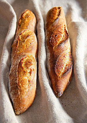 „Der große Lafer Backen“: Die Baguettes im Vergleich: rechts Lafers Rezeptur, links meine veränderte Fassung. Die beiden Baguettes weisen bereits im nicht angeschnittenen Zustand deutliche Unterschiede auf. Das Baguette von Lafer ist deutlich kleiner geblieben und die Schnitte sind nicht so rustikal aufgebrochen.