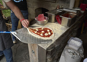 Ein mit Tomatensauce und Mozzarella belegter Pizzateigling liegt zur Hälfte auf der mit Hartweizengrieß bestreuten Arbeitsfläche, zur anderen Hälfte auf einem metallenen Pizzaschieber.