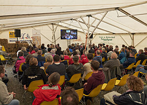 Eng aneinandergereiht sitzen Zuschauer in einem Zelt vor einem großen Bildschirm, der die Vorträge des Symposiums zeigt.