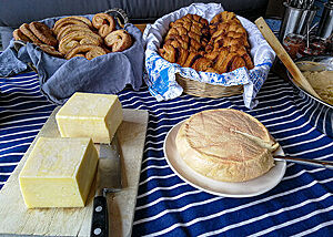 Auf einem Tisch stehen zwei Körbe mit Schweinsohren und Franzbrötchen, ein Holzbrett mit Butter und ein Teller mit Käse.