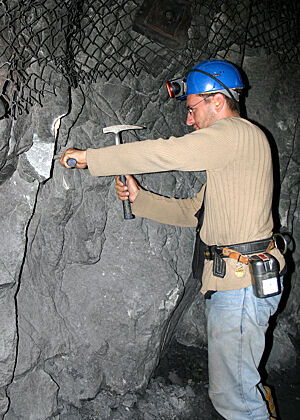 Ein junger Mann in Bergmannausrüstung schlägt Gesteins-Proben aus einer Stollenwand.