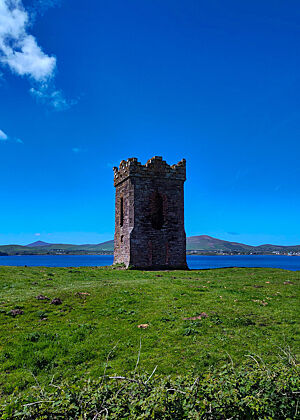 Hussey's Folly – Dieser Turm am Eingang zur Bucht von Dingle wurde als eine Art Büro gebaut, um Zoll von einfahrenden Schiffen kassieren zu können. Ein steinerner Turm auf grüner Wiese, im Hintergrund die Bucht von Dingle mit strahlend blauem Wasser.
