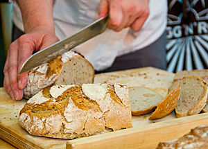 Eine männliche Person im Bildhintergrund hält das Brot mit seiner Rechten, mit der Linken schneidet er eine Scheibe ab. Im Vordergrund ein halber Laib frischgebackenen Brots.