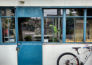 Blick auf eine weiße Hauswand mit blauer Tür und blauen Fenstern. Davor steht ein Fahrrad.