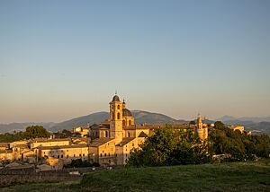 Nach unserem Rundgang ein letzter Blick auf Urbino im Sonnenuntergang. Die Stadt Urbino strahlt im letzten Abendlicht. In der Mitte erhebt sich ein Kirchturm und im Hintergrund sieht man grüne Hügel.
