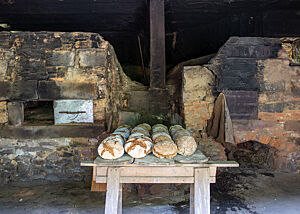 Zwei Öfen stehen nebeneinander, während in der Mitte davor ein mit fertigen Brotlaiben beladener Holztisch steht. 