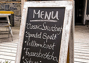 Backen für „Bread & Dance“. Ein Aufsteller mit Kreidetafel trägt die Aufschrift: „Menu: Classic Sourdough, Sprouted Spelt, Vollkornbrot, Franzbrötchen, Schweinsöhrchen“.