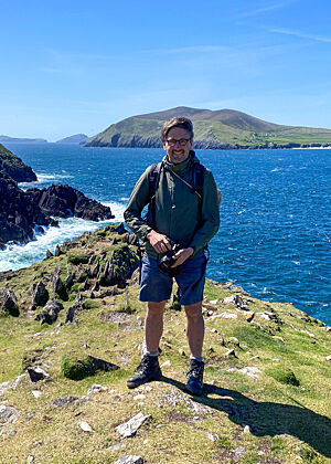 Lutz Geißler steht in Wander-Outfit mit Kamera auf den Sandsteinen des Dunmore Head, umgeben von azurblauem Wasser.