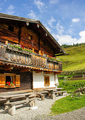 Roswithas Hütte. Die Hütte von Roswitha Huber, mit Bänken und Tischen vor der Tür, in den Bergen, vor grünen Wiesen und blauem, leichte bewölktem Himmel.