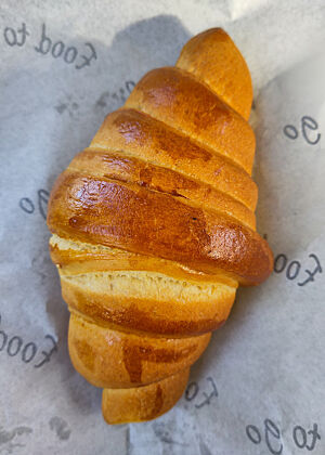 Das „Plain Croissant“ von My Patrik's Bakery – eigentlich eine Art Hörnchen aus Feinteig, jedenfalls nicht touriert wie ein Croissant (also ohne Butterschichten). Ein goldbraun gebackenes „Croissant“ auf Einschlagpapier.
