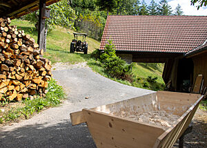 Im Vordergrund steht eine eckige Backmulde aus hellem Holz, zur Hälfte gefüllt mit Brotteig. Im Hintergrund stapelt sich unter einem Ziegeldach Holz zum Trocknen.