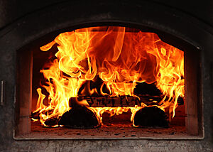 Durch die Öffnung des Holzbackofens sind brennende Holzscheite und Flammen zu sehen.