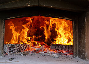 Anheizen des Holzofens für das Schwarzwaldbrot in Mittelitalien. Durch die geöffnete Ofentür ist ein loderndes Feuer zu sehen.