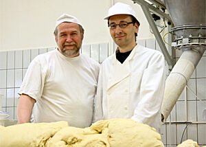 Portrait von Bäcker Süpke und Lutz: Beide stehen in typischer Bäcker-Arbeitskleidung hinter einer großem Berg von frisch geknetetem Teig
