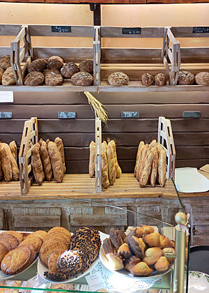 Blick auf die Auslage von Petit Delice mit Baguettes und Broten. Eine Backwarenauslage in Holzoptik mit vielen Baguettes und Broten im Hintergrund. Im Vordergrund werden Madeleines und anderes Süßgebäck präsentiert.