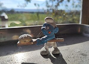 Backen im Land der Schlümpfe Eine Schlumpffigur mit einem Brotschieber und einem Brot darauf in den Händen steht auf einer Metallschale in der Fensterbank, durch die die Sonne ins Backhaus scheint.