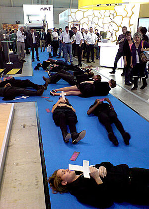 Schwarz gekleidete Studenten der Uni Hohenheim liegen mit weißen Kreuzen in der Hand und fiktiven Pappbroten auf dem blauen Messeteppich.