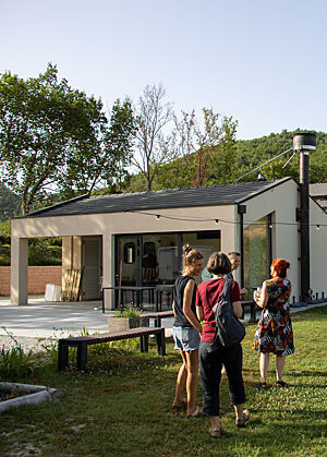 Eva und Giuditta im Gespräch mit Sabine und Christina. Im Hintergrund die Backstube der Bäckerei Il Gentil Verde in Acqualagna. Eva, Giuditta, Sabine und Christina stehen vor dem modernen Gebäude mit weißen Wänden und anthrazitfarbenem Dach, in dem sich die Backstube befindet. Außen führt ein dicker Schornstein den Rauch nach oben.