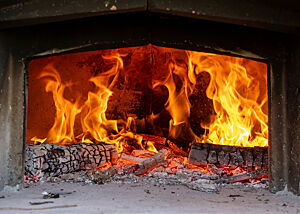 Anheizen des Holzofens für das Schwarzwaldbrot in Mittelitalien. Durch die geöffnete Ofentür ist ein loderndes Feuer zu sehen.