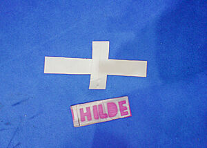 Ein weißes Kreuz und fiktives Pappbrot mit der Aufschrift „Hilde“ liegen auf dem blauen Messeteppich.