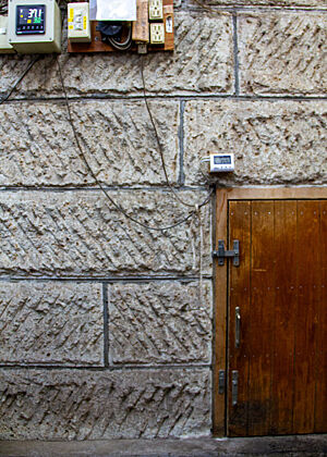 Eine Wand aus Steinen mit Elektronik, deren Kabel zu einer braunen Holztür führen.